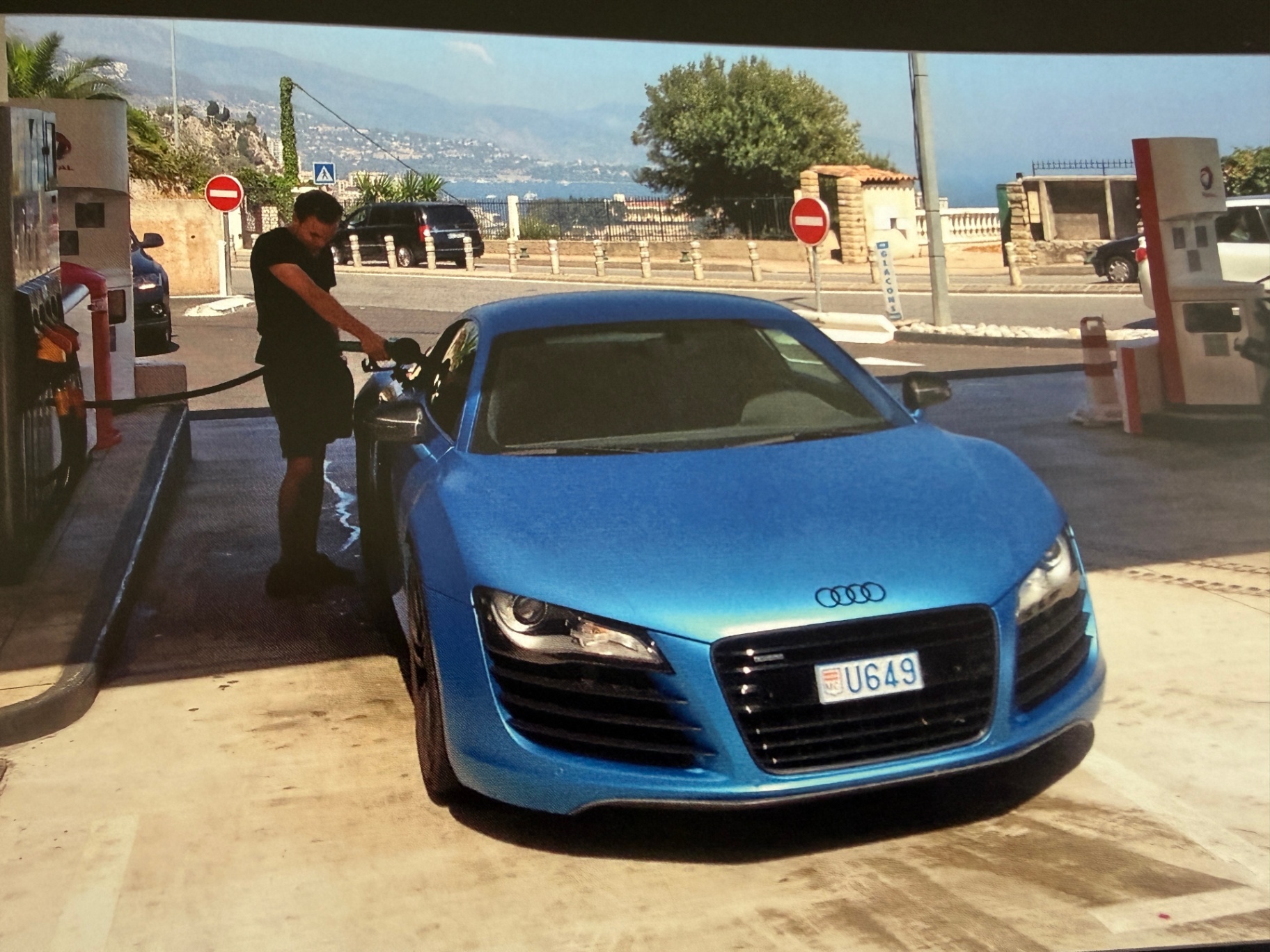 Blauwe Audi R8 met Monaco-kenteken bij een tankstation.